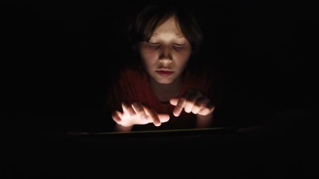 Dolly shot caucasian girl lying in bed playing tablet in social internet in dark light under blacket. Childhood, communication and technology concept