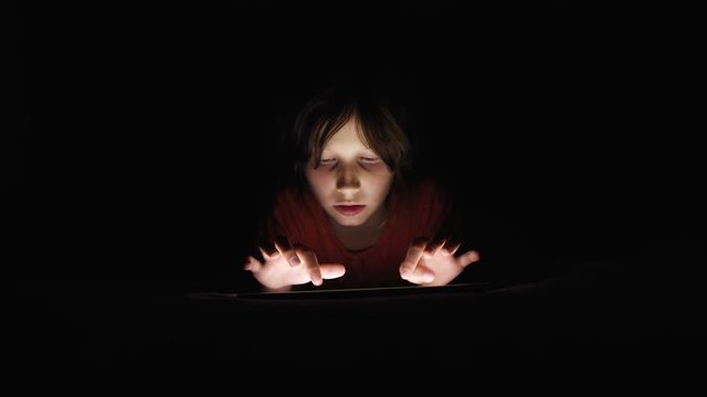 Dolly shot caucasian girl lying in bed playing tablet in social internet in dark light under blacket. Childhood, communication and technology concept