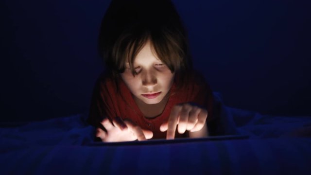 Dolly shot caucasian girl lying in bed playing tablet in social internet in dark blue light under blacket. Childhood, communication and technology concept