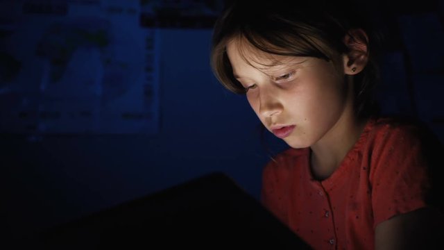 Dolly shot caucasian girl lying in bed playing tablet in social internet in dark blue light under blacket. Childhood, communication and technology concept