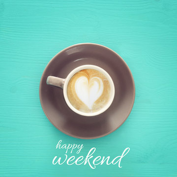 image of coffe cup with foam of heart shape over wooden blue background and text: HAPPY WEEKEND.