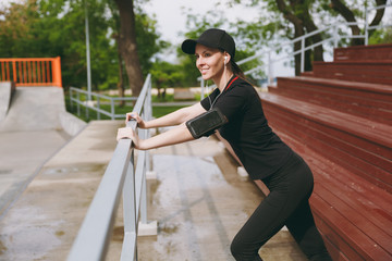 Young smiling athletic beautiful brunette girl in black uniform, cap with headphones listening music doing sport stretching exercises warming-up in city park outdoors. Fitness, healthy lifestyle.