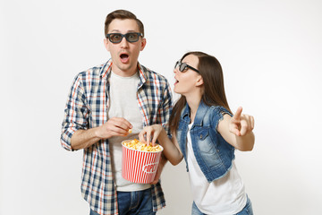 Young shocked couple, woman and man in 3d glasses watching movie film on date, holding bucket of popcorn, stealing popcorn, pointing index finger isolated on white background. Emotions in cinema.