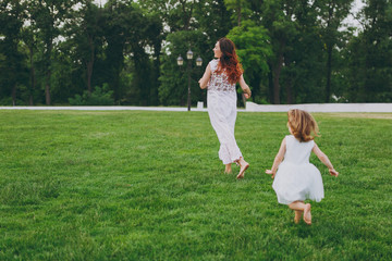 Fototapeta premium Little cute child baby girl running catching up have fun with joyful woman in light dress on green grass in park. Mother, little kid daughter. Mother's Day, love family, parenthood, childhood concept.