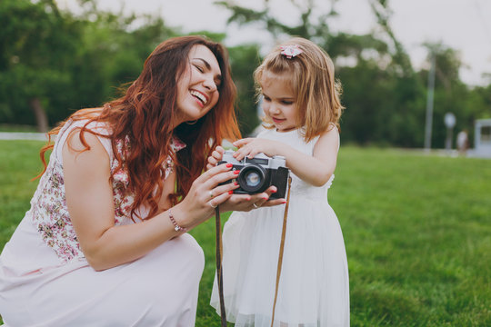 Laughing woman in light dress and little pretty child baby girl holding retro vintage photo camera in green park. Mother, little kid daughter. Mother's Day, love family, parenthood, childhood concept.