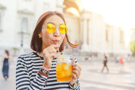 Happy female hipster student drinks a cool lemonade through a straw in a summer outdoor cafe