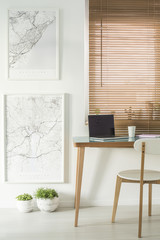 Maps on the wall next to a desk with a laptop and chair standing by the window with blinds in a...