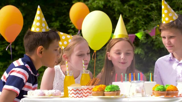 holidays, childhood and celebration concept - happy kids blowing out candles on birthday cake sitting at table at summer garden party
