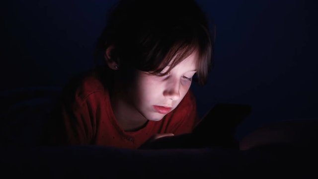 Dolly shot caucasian girl lying in bed playing tablet in social internet in dark blue light under blacket. Childhood, communication and technology concept