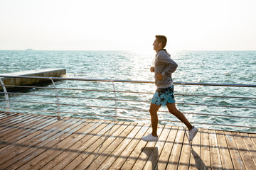 Profile of young motivated sportsman, jogging on quay, near the ocean. Full length. Dressed in sport shorts and jacket. Outdoors.