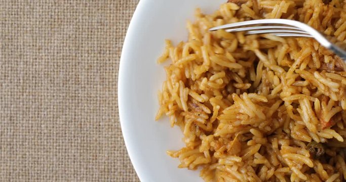 Top close view of using a fork to get a forkful of spicy rice on a white plate atop a burlap tablecloth.
