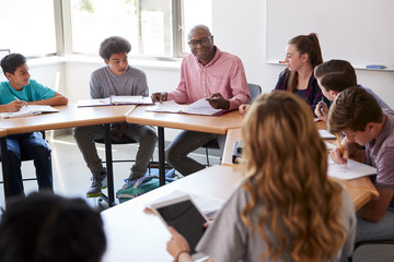 Male High School Tutor Sitting With Students At Desk In Class