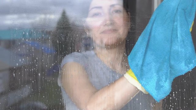 Beautiful woman washes window with a blue rag and cleanser, slow motion