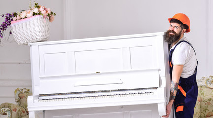 Loader moves piano instrument. Delivery service concept. Man with beard, worker in overalls and helmet lifts up piano, white background. Courier delivers furniture in case of move out, relocation. © be free
