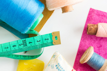 sewing, patchwork and tailoring concept - closeup on measuring meter, blue and pink threads in bobbins and purple and mustard color patches on white background, top view, flat lay.