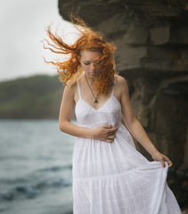 Fototapeta na wymiar Redhead woman in white dress standing on beach and looking to somewhere.