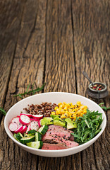 Healthy dinner. Buddha bowl lunch with grilled beef steak and quinoa, corn, avocado, cucumber and arugula on wooden background. Meat salad.