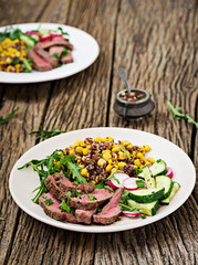 Healthy dinner. Bowl lunch with grilled beef steak and quinoa, corn, cucumber, radish and arugula on wooden background. Meat salad.