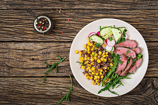 Healthy dinner. Bowl lunch with grilled beef steak and quinoa, corn, cucumber, radish and arugula on wooden background. Meat salad. Flat lay. Top view