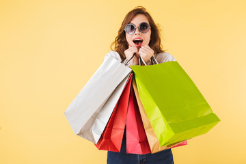 Portrait of young surprised lady in sunglasses standing with colorful shopping bags in hands and...
