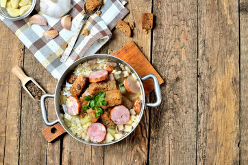 Top view of old saucepan with garlic soup, fresh garlic bulbs, cloves, colored pepper, wooden spoon, croutons, lable and a towel on a rustic wooden table