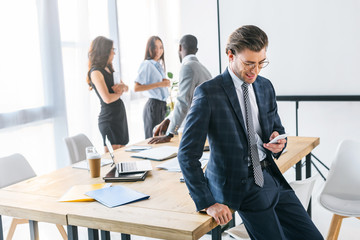 selective focus of caucasian businessman using smartphone and multiracial colleagues behind in office