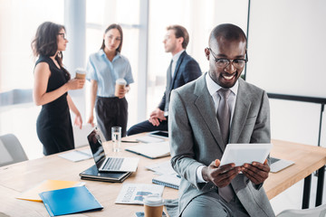 selective focus of smiling african american businessman using tablet and colleagues at workplace in office