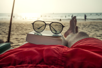 sunglasses lying on a book of person on the sandy beach in front of water and sunny sky with clouds