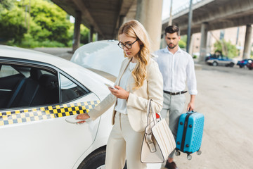 stylish young woman woman using smartphone while man putting suitcase in trunk of taxi