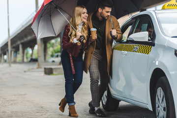smiling young couple with umbrellas looking at taxi car - obrazy, fototapety, plakaty