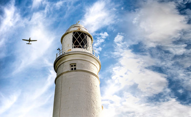 Coastal lighthouse with plane flying above under summer sky.