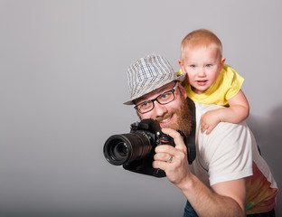 A photographer with a child in his arms takes an enthusiastic photograph against a gray background in the studio. The concept of "Children of Photography is not a hindrance"
