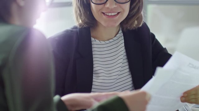 Tilt up of young beautiful businesswoman in eyeglasses discussing documents with female colleague at office table