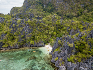 Aerial drone view of boats around a tiny sandy beach on a tropical island surrounded by towering limestone cliffs