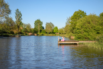 Fototapeta na wymiar Ein Schulmädchen sitzt auf dem Steg an einem See und angelt
