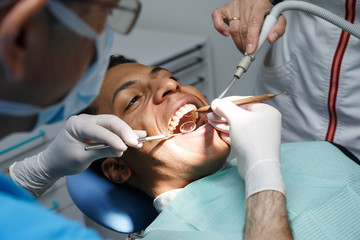 Dentist examining oral cavity of young African-American man working in in dental clinic with assistant. 