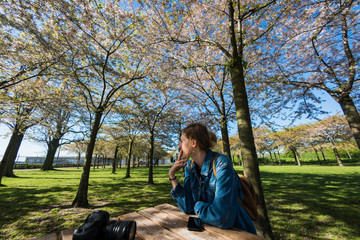 young woman smoking cigarette while sitting at wooden table with camera and smartphone in beautiful park with blossoming trees, copenhagen, denmark