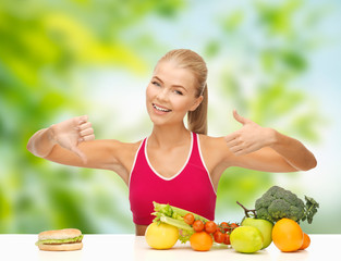 diet, junk food and eating concept - happy smiling woman with vegetables and hamburger showing thumbs up and down sign over green natural background