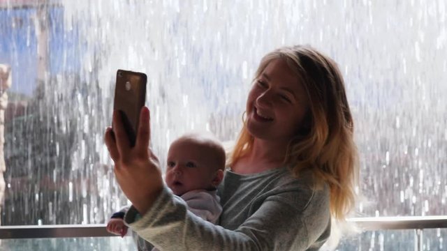 weekend, young mother with baby in arms doing selfie photo using mobile phone on a background of water wall