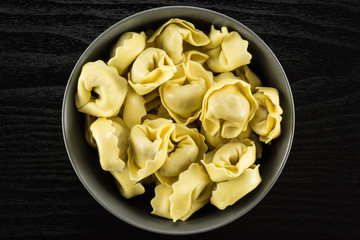 Raw tortellini pasta in a grey ceramic bowl flatlay on black wood background Italian traditional dumpling.