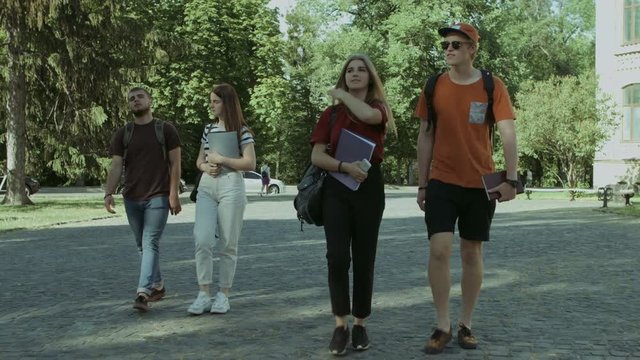 Cheerful student friends with textbooks and backpacks going to university. Group of college students walking in university campus to study. Steadicam stabilized shot.