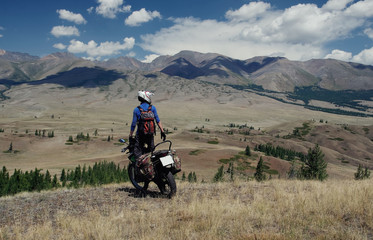 Motorcycle traveler man in helmet with suitcases standing on extreme rocky road in a mountain valley in cloudy weather on the background of endless steppe
