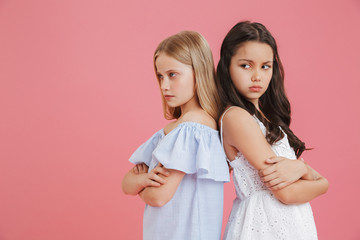 Picture of offended brunette and blonde girls 8-10 years old wearing dresses standing back to back with arms crossed and expressing argument, isolated over pink background