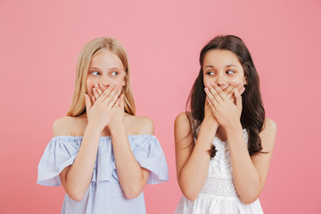 Image of two cute girls 8-10 wearing dresses covering mouthes with hands and looking to each other,...