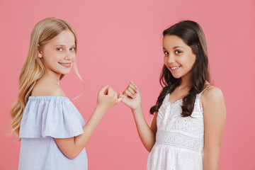Photo of lovely little girls 8-10 years old wearing dresses hook each other's little fingers in conciliation or friendship, isolated over pink background