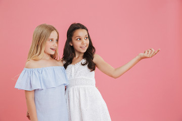 Image of two excited girls 8-10 years old wearing dresses looking aside and pointing finger at copyspace, isolated over pink background