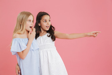 Image of two surprised and happy girls 8-10 years old wearing dresses looking aside and pointing finger at copyspace, isolated over pink background
