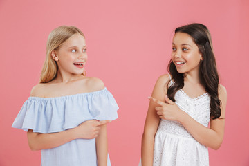 Picture of happy brunette and blonde girls 8-10 years old wearing dresses smiling and pointing finger at each other, isolated over pink background