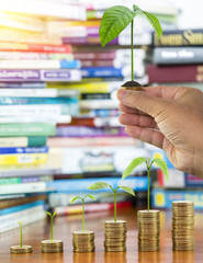 Man hand holding tree growing and soil on money coins stack arranged as a graph with blurred pile of book as background, concept of educational advancement and saving money