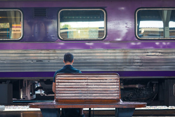 back of a man waiting on a bench at railway station with purple train in background during day
